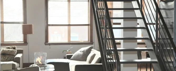 Living room with stairs, couch, windows, and exposed ductwork. Sunlight streams in.