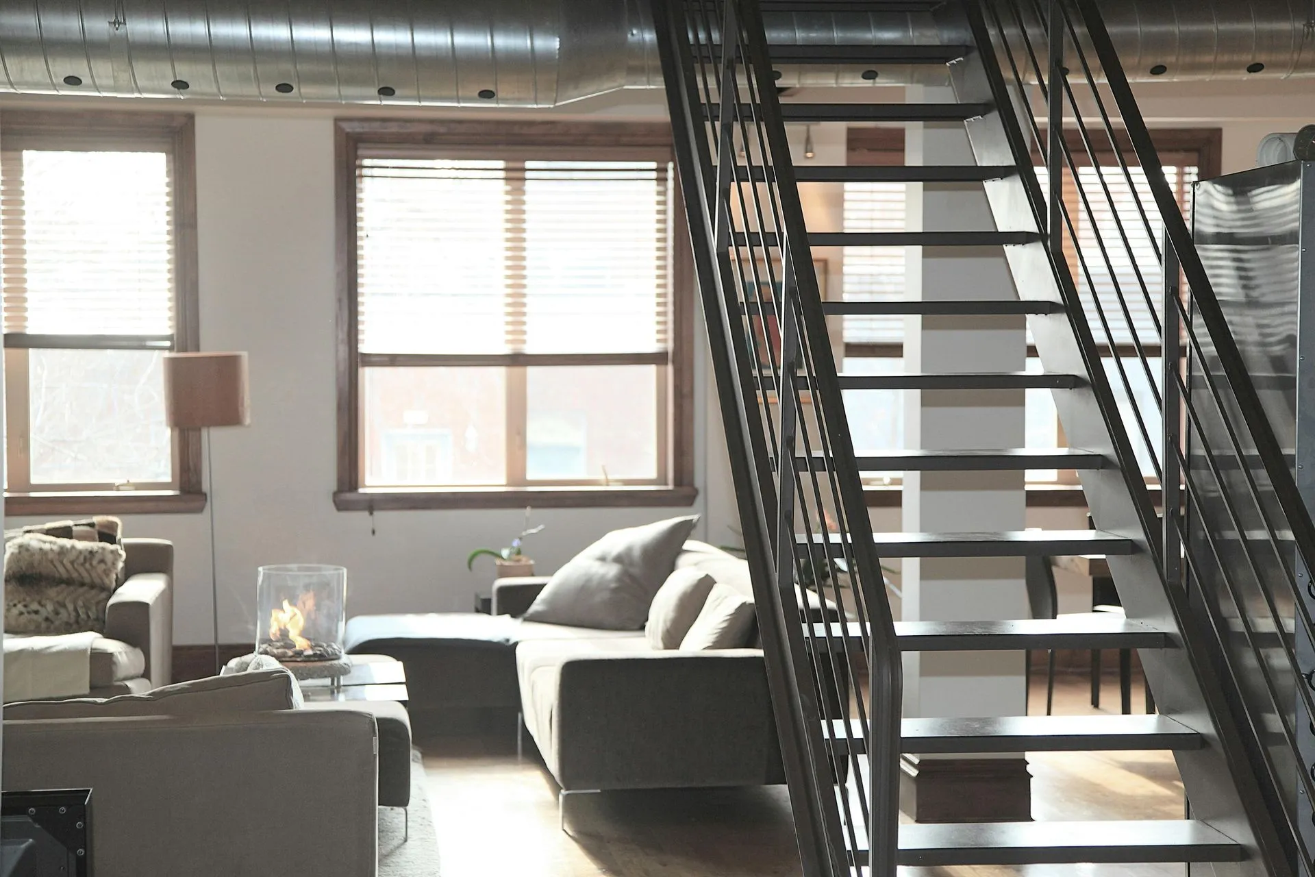 Living room with stairs, couch, windows, and exposed ductwork. Sunlight streams in.